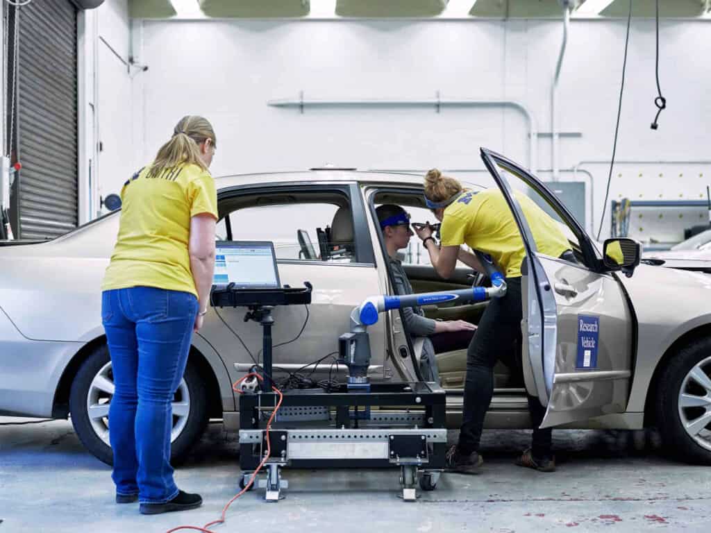 Two researchers conducting ergonomic research on research subject seated in the driver's seat of a vehicle.