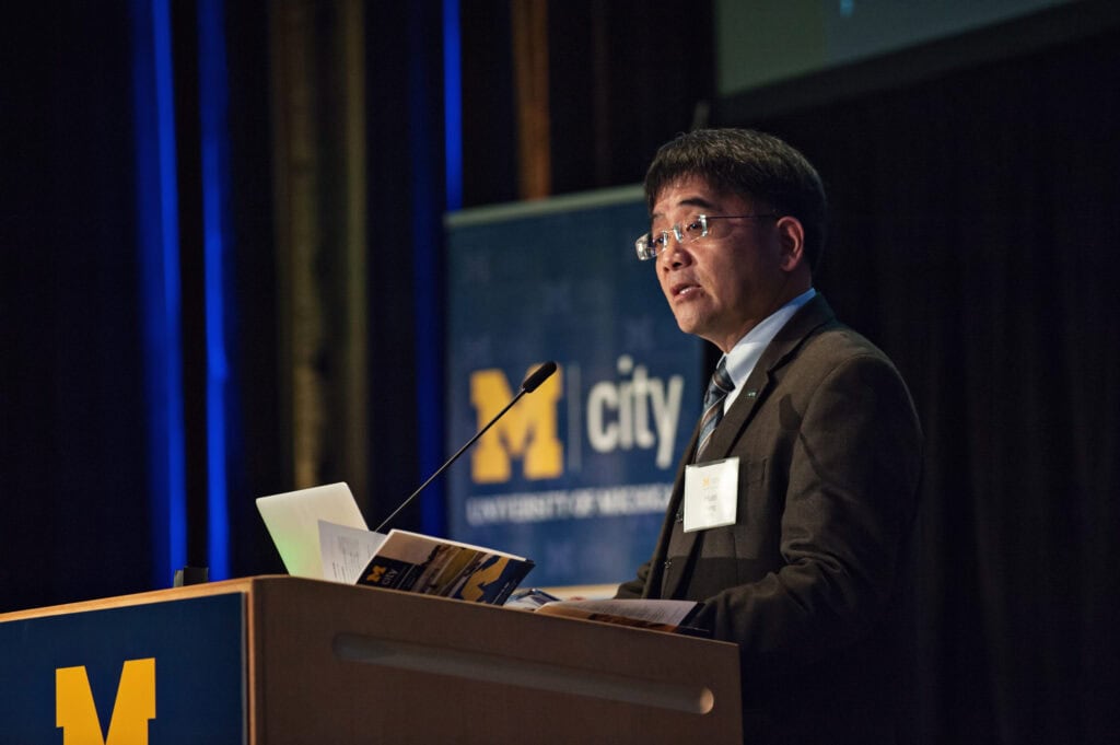 Professor Huei Peng stands at podium with Mcity banner behind his right shoulder. Banner is blue with white print. Professor Peng has black hair and wears glasses. He is wearing brown jacket, white shirt and tie.